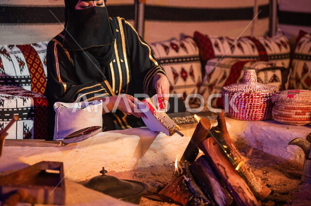 Using the popular pump to light a fire, following authentic traditional Saudi customs, preparing Arabic coffee inside a heritage tent, a Saudi Gulf Arab woman wearing a burqa and black abaya and using a firewood blower, enjoying preparing the stove to roast coffee beans