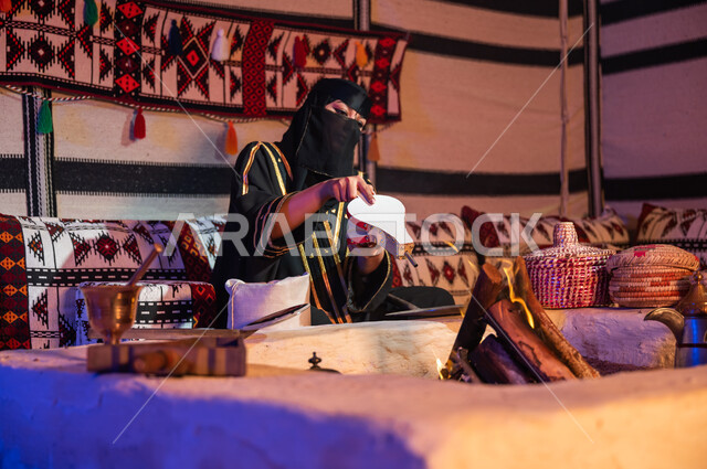 Preparing Arabic coffee inside a heritage tent, following authentic traditional Saudi customs, a Saudi Gulf Arab woman wearing a burqa and black abaya and using a firewood blower, using the popular pump to light a fire, enjoying preparing the stove to roast coffee beans