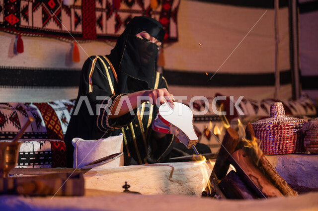 اتباع العادات السعودية التقليدية الأصيلة, تحضير القهوة العربية داخل خيمة تراثية, امرأة عربية خليجية سعودية ترتدي البرقع والعباءة السوداء وتقوم باستخدام منفاخ الحطب, استعمال المضخة الشعبية لإشعال النار, الاستمتاع بتجهيز الموقد لتحميص حبوب البن