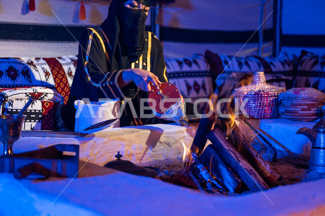 الجلوس داخل خيمة تراثية شعبية, اتباع العادات والتقاليد السعودية في تحضير القهوة العربية, امرأة عربية خليجية سعودية ترتدي البرقع والعباءة السوداء وتقوم باستخدام منفاخ الحطب, استعمال المضخة الشعبية لإشعال النار, تجهيز الموقد لتحميص حبوب البن