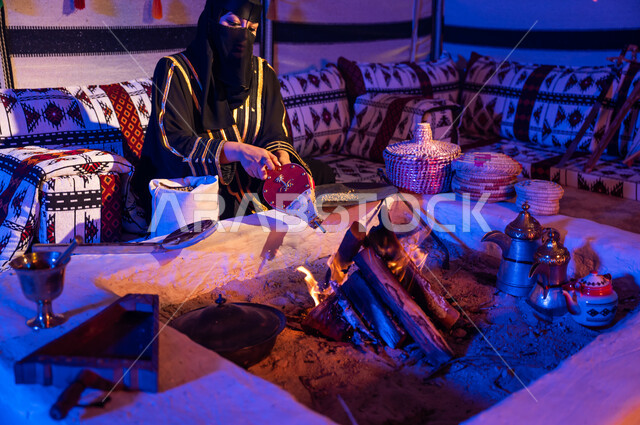 الاستمتاع بالجلسات الشعبية داخل خيمة تراثية, اتباع العادات والتقاليد السعودية في تحضير القهوة العربية, امرأة عربية خليجية سعودية ترتدي البرقع والعباءة السوداء وتقوم باستخدام منفاخ الحطب, استعمال المضخة الشعبية لإشعال النار, تجهيز الموقد لتحميص حبوب البن