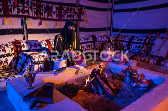 تجهيز الموقد لتحميص حبوب البن, اتباع العادات والتقاليد السعودية في تحضير القهوة العربية, امرأة عربية خليجية سعودية ترتدي البرقع والعباءة السوداء وتقوم باستخدام منفاخ الحطب, استعمال المضخة الشعبية لإشعال النار, الاستمتاع بالجلسات الشعبية داخل خيمة تراثية