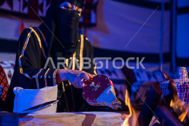 العادات والتقاليد السعودية في تحضير القهوة, صورة مقربة لامرأة عربية خليجية سعودية ترتدي البرقع والعباءة السوداء وتقوم باستخدام منفاخ الحطب, استعمال المضخة الشعبية لإشعال النار, تجهيز الموقد لتحميص حبوب البن, الاستمتاع بالجلسات الشعبية داخل خيمة تراثية