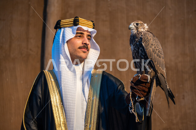 Looking at the falcon with gestures of strength and challenge, adhering to the Kingdom’s civilizational and cultural heritage, a close-up of a Saudi Gulf Arab man wearing a ghutra, a bisht, and an aqal holding a falcon bird on his hand, the passion of the Saudis for raising birds of prey, the relationship with falcons, the founding of the kingdom February 22, founding day 1727