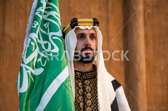إحياء يوم العلم 11 مارس, ارتداء الملابس الشعبية للاحتفال باليوم الوطني 23 سبتمبر, صورة مقربة لرجل عربي خليجي سعودي يرتدي الدقلة والغترة والعقال الذهبي ويحمل بيده العلم السعودي وينظر إلى شيء ما, ذكرى تأسيس الدولة السعودية الأولى 1727, الافتخار بالهوية الوطنية