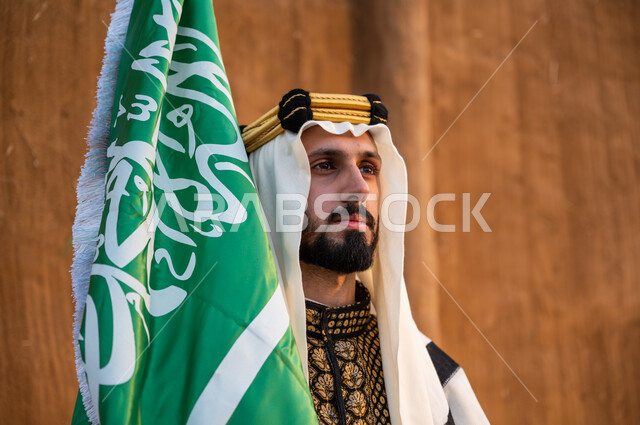 الافتخار بالهوية الوطنية, ارتداء الملابس الشعبية للاحتفال باليوم الوطني 23 سبتمبر, إحياء يوم العلم 11 مارس, صورة مقربة لرجل عربي خليجي سعودي يرتدي الدقلة والغترة والعقال الذهبي ويحمل بيده العلم السعودي وينظر إلى شيء ما, ذكرى تأسيس الدولة السعودية الأولى 1727