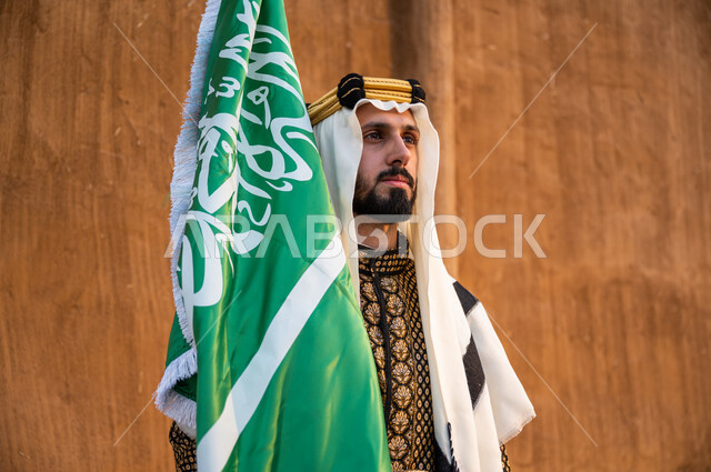 ذكرى تأسيس الدولة السعودية الأولى 1727, ارتداء الملابس الشعبية للاحتفال باليوم الوطني 23 سبتمبر, إحياء يوم العلم 11 مارس, صورة مقربة لرجل عربي خليجي سعودي يرتدي الدقلة والغترة والعقال الذهبي ويحمل بيده العلم السعودي وينظر إلى شيء ما, الافتخار بالهوية الوطنية