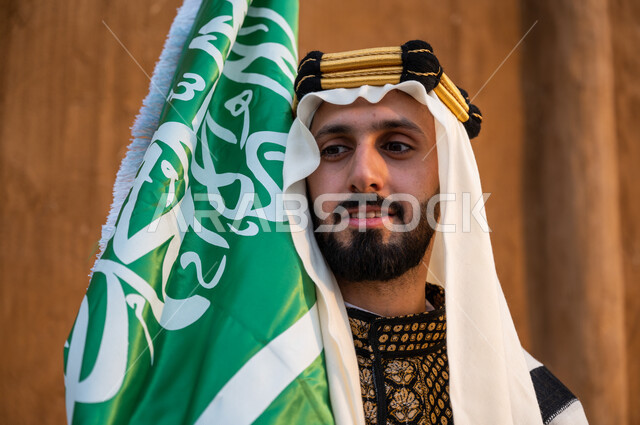 ارتداء الملابس الشعبية للاحتفال باليوم الوطني 23 سبتمبر, إحياء يوم العلم 11 مارس, صورة مقربة لرجل عربي خليجي سعودي يرتدي الدقلة والغترة والعقال الذهبي ويحمل بيده العلم السعودي وينظر إلى شيء ما, ذكرى تأسيس الدولة السعودية الأولى 1727, الافتخار بالهوية الوطنية
