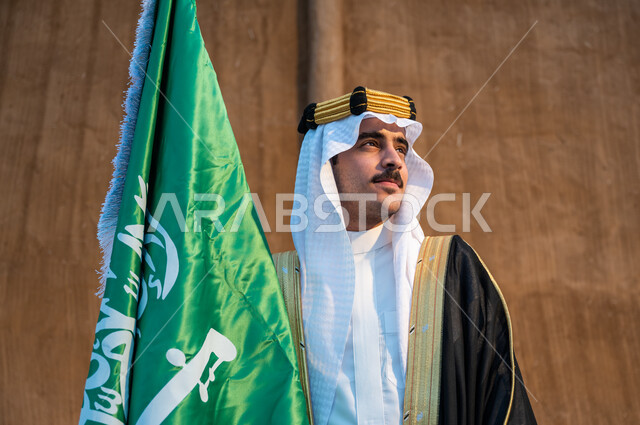 الفخر بالهوية الوطنية السعودية، الوقوف باعتزاز والنظر الى شيء ما, صورة مقربة لرجل عربي خليجي سعودي يرتدي البشت والعقال والغترة يحمل بيده علم المملكة، ذكرى يوم العلم 11 مارس، اليوم الوطني السعودي 23 سبتمبر، تأسيس الدولة السعودية 1727 م، خلفية جدار طيني