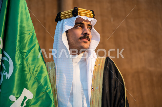 Standing proudly and looking at something, pride in Saudi national identity, a close-up photo of a Saudi Gulf Arab man wearing a bisht, aqal and a ghutra holding the Kingdom’s flag in his hand, commemoration of Flag Day March 11, Saudi National Day September 23, founding of the Saudi state 1727 AD, background of a mud wall
