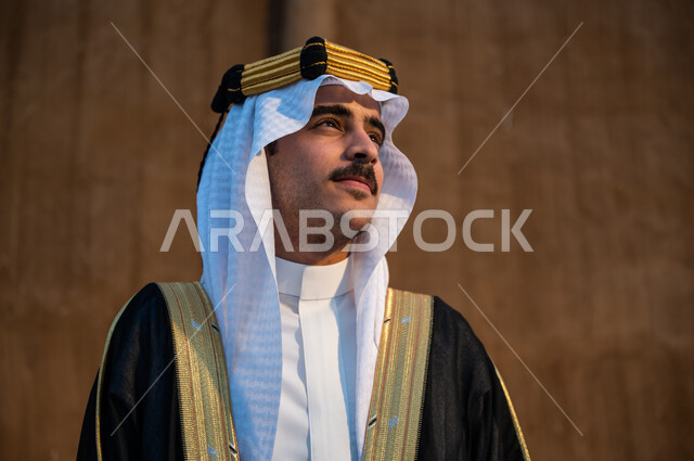 ارتداء أزياء المناسبات والأعياد الوطنية, النظر والتأمل وإظهار مشاعر حب الوطن, صورة مقربة لرجل عربي خليجي سعودي يرتدي العقال والبشت والغترة ينظر إلى شيء ما, استذكار يوم تأسيس الدولة السعودية الأولى 1727م,  ذكرى يوم التأسيس في 22 فبراير, خلفية جدار طيني