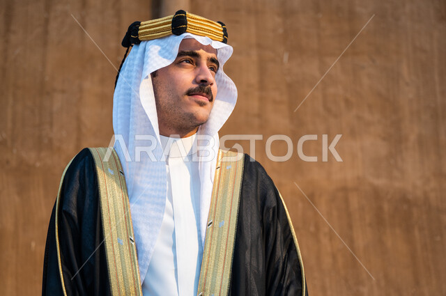 النظر والتأمل وإظهار مشاعر حب الوطن, ارتداء أزياء المناسبات والأعياد الوطنية, صورة مقربة لرجل عربي خليجي سعودي يرتدي العقال والبشت والغترة ينظر إلى شيء ما, استذكار يوم تأسيس الدولة السعودية الأولى 1727م,  ذكرى يوم التأسيس في 22 فبراير, خلفية جدار طيني