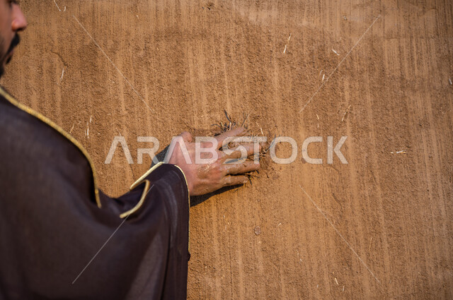 التمسك والتعلق بالهوية الوطنية, إظهار مشاعر الحب والولاء للدولة, صورة مقربة من الخلف لرجل عربي خليجي سعودي يرتدي البشت الأسود يقوم بجمع حفنة تراب, ذكرى يوم التأسيس السعودي في 22 فبراير، تأسيس الدولة السعودية الأولى 1727م، خلفية جدار طيني