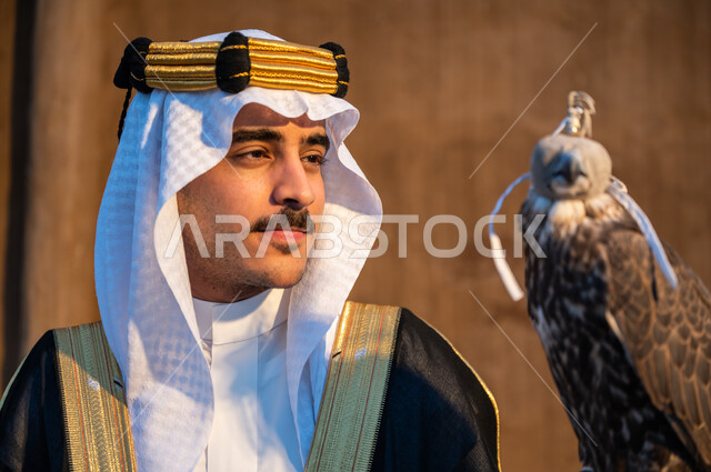 مكانة الصقور في التاريخ السعودي وعلاقتها بيوم التأسيس 1727م, تأسيس الدولة السعودية الأولى 22 فبراير, صورة مقربة لرجل عربي خليجي سعودي يرتدي الغترة والبشت والعقال الذهبي يحمل على يده طائر الصقر المغطى بخوذة الرأس الجلدية وينظر اليه بحب