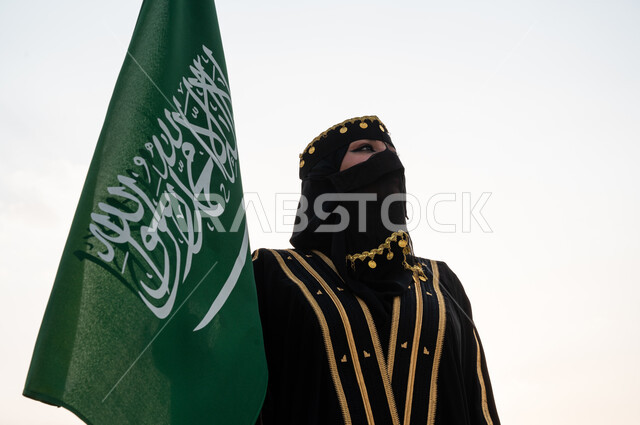 ذكرى يوم التأسيس 22 فبراير, صورة مقربة لسيدة عربية خليجية سعودية ترتدي العباءة السوداء والبرقع وتقف بجانب علم المملكة, يوم العلم السعودي 11 مارس, الاحتفال باليوم الوطني 23 سبتمبر, التفاخر بالإنتماء للوطن, ارتداء الأزياء الشعبية التقليدية في المناسبات الوطنية