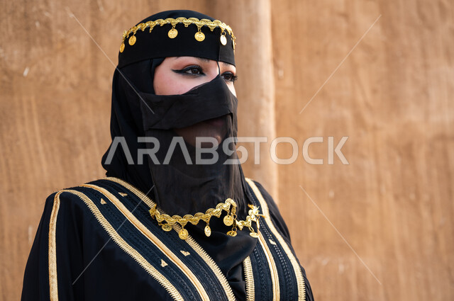 الوقوف بفخر واعتزاز، المناسبات والاعياد الوطنية، ارتداء أزياء أهل المنطقة الوسطى، صورة مقربة لإمرأة سعودية ترتدي العباءة و البرقع  تتأمل وتنظر إلى شيء ما، ذكرى يوم التأسيس السعودي في 22 فبراير، تأسيس الدولة السعودية الأولى 1727 م، خلفية جدار مبني من الطين