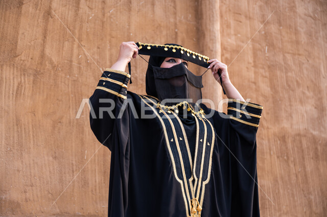 التجهز للاحتفال بيوم التأسيس 22 فبراير, ارتداء الملابس الشعبية السعودية, صورة مقربة لإمرأة عربية خليجية سعودية ترتدي العباءة السوداء وتقوم بلبس البرقع, الاعتزاز بذكرى تأسيس المملكة 1727, الأنوثة والجمال البدوي للنساء السعوديات, الوقوف باستقامة و فخر