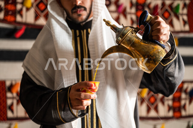 كرم الضيافة وحسن الإستقبال وفق العادات والتقاليد الأصيلة, الاستمتاع بشرب البن العربي في الخيمة الشعبية، التمسك بالقيم الكريمة المتوارثة من الأجداد ، صورة مقربة لرجل عربي خليجي سعودي يرتدي الدقلة والغترة و يقوم بصب القهوة في الفنجان