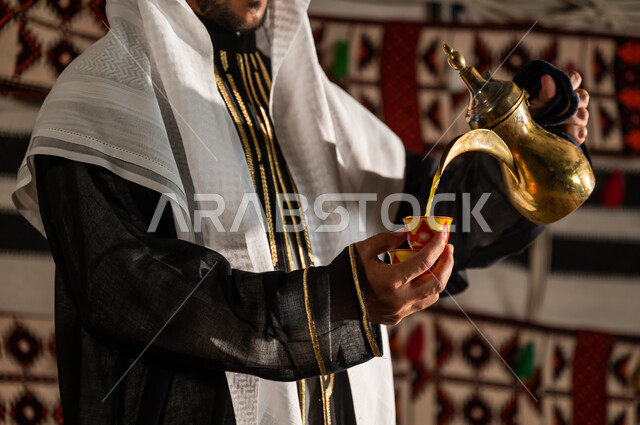 شرب البن العربي في الخيمة الشعبية، التمسك بالقيم الكريمة المتوارثة من الأجداد ، صورة مقربة لرجل عربي خليجي سعودي يرتدي الدقلة والغترة و يقوم بصب القهوة في الفنجان، أساليب عريقة في كرم الضيافة، مفهوم حسن الإستقبال وفق العادات والتقاليد الأصيلة