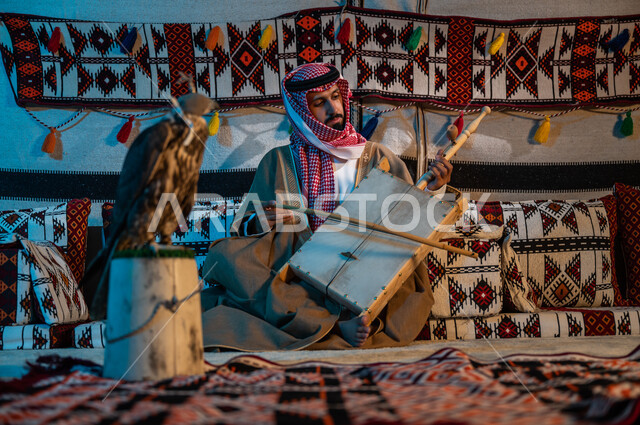 Traditional Saudi sessions in heritage tents, following the ancestral approach in raising falcons, a Saudi Gulf Arab man wearing a shemagh and a bisht playing the rabab instrument, popular artistic musical talents, a hobby of playing string instruments, a close-up of a falcon bird covered with a leather head helmet.