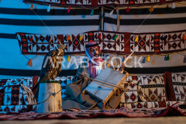 الجلوس في خيمة تراثية والاستمتاع بالعزف, حب امتلاك وتربية الطيور الجارحة، المحافظة على الموروث الثقافي الفني، رجل عربي خليجي سعودي يرتدي الشماغ والبشت يقوم بالعزف على آلة الربابة، الآلات الوترية الموسيقية الشعبية، طائر الصقر المغطى بخوذة الرأس