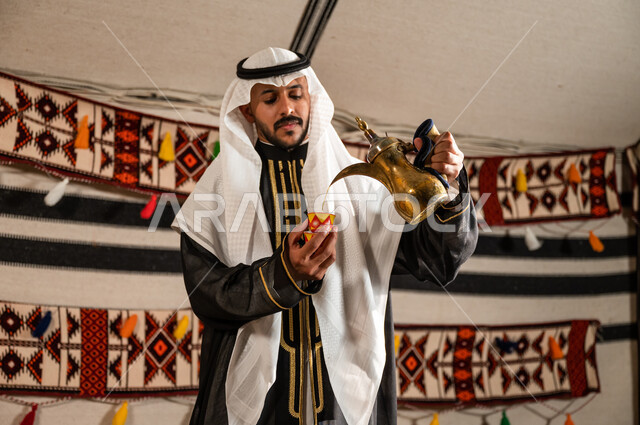 Providing local products to guests in traditional folk tents, following the path of our ancestors in honoring the guest, a close-up picture of a Saudi Gulf Arab man wearing a dahla and ghutra and pouring coffee from a copper pot, authentic customs and traditions in serving coffee, inheriting good reception and generosity.