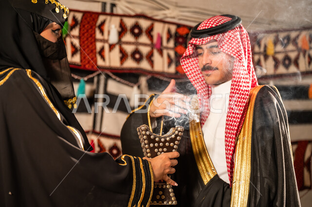 ارتداء أزياء أهل المنطقة الشرقية، العناية بمظهر الزوج الخارجي من خلال التبخير والتطيب، امرأة عربية خليجية سعودية ترتدي العباية السوداء والبرقع وتمسك حامل البخور (المبخرة) وتقوم بتعطير زوجها، مفهوم الترابط الاسري والحياة السعيدة, روائح عطرية وطنية مميزة