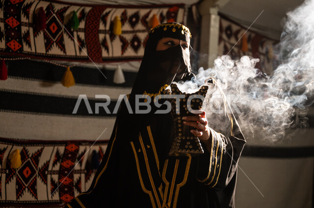 اتباع عادات وتقاليد المملكة الأصيلة، التبخر باستخدام خشب العود والمبخرة العربية في الأعياد والمناسبات الوطنية، صورة مقربة لإمرأة عربية خليجية سعودية ترتدي البرقع و العباءة السوداء تقوم بالتعطر والتطيب  بالبخور العربي داخل خيمة تراثية