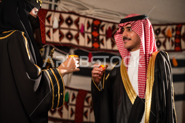 ارتداء البشت والأزياء الشعبية التقليدية، تقديم منتجات وطنية محلية, العلاقات الزوجية السعودية السعيدة، سيدة عربية خليجية سعودية ترتدي العباءة السوداء والبرقع وتقوم بمنح القهوة لزوجها، الإستمتاع بشرب القهوة العربية في خيمة تراثية