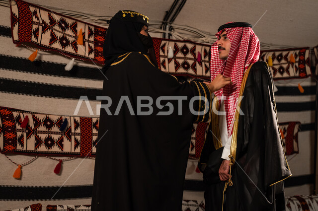 مفهوم العشرة الطيبة و المودة بين الزوجين, العلاقات الزوجية السعيدة , اهتمام المرأة بمظهر زوجها، زوجة عربية سعودية خليجية ترتدي البرقع والعباءة السوداء تقوم بتعديل لباس الزوج، ارتداء الأزياء الشعبية الوطنية