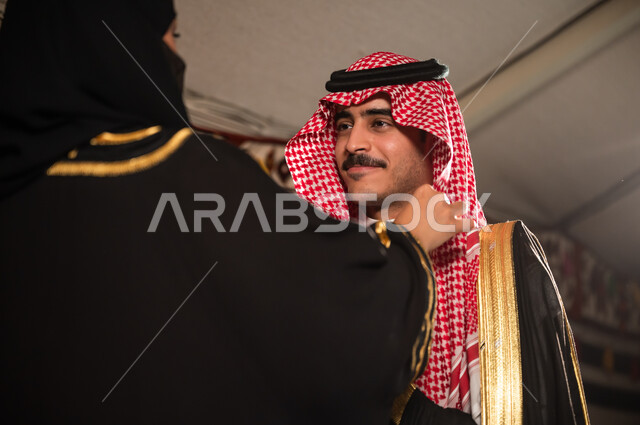 ارتداء الأزياء الشعبية الوطنية، العلاقات الزوجية السعيدة , اهتمام المرأة بمظهر زوجها، زوجة عربية سعودية خليجية ترتدي البرقع والعباءة السوداء تقوم بتعديل لباس الزوج، مفهوم الاهتمام و المودة بين الزوجين