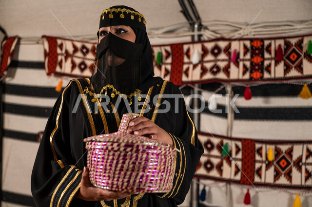 أصالة العادات والتقاليد السعودية, الوقوف داخل خيمة تراثية شعبية والنظر إلى شيء ما, حسن الاستقبال والضيافة, امرأة عربية خليجية سعودية ترتدي البرقع والعباءة تحمل بيدها سلة من القش,  القيم و المبادئ التقليدية المتوارثة في الكرم