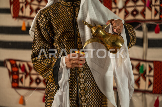 عادات وتقاليد أصيلة في تقديم القهوة, السير على نهج الأجداد في إكرام الضيف, صورة مقربة لرجل عربي خليجي سعودي يرتدي الدقلة والغترة ويقوم بصب القهوة من الدلة النحاسية, توارث حسن الاستقبال والكرم, تقديم المنتجات المحلية للضيوف في الخيام الشعبية التراثية