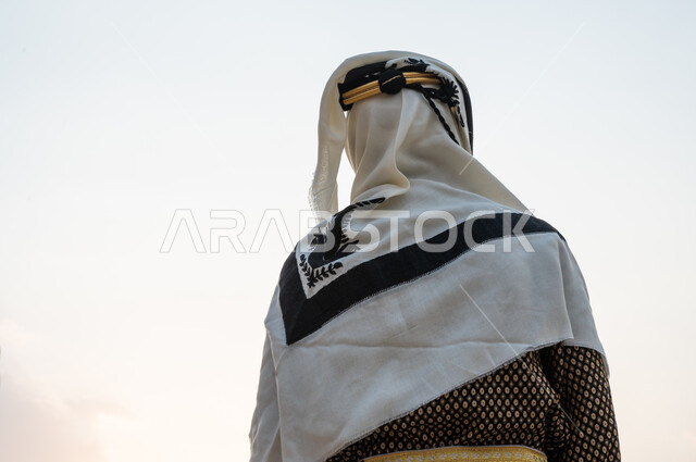 الوقوف والتأمل بافتخار واعتزاز, صورة مقربة من الخلف لرجل عربي خليجي سعودي يرتدي الدقلة والعقال الذهبي و ينظر إلى السماء, ذكرى تأسيس الدولة السعودية الأولى 1727م, ارتداء الأزياء التراثية الشعبية في يوم التأسيس 22 فبراير, حب الوطن والانتماء إليه, خلفية السماء الصافية