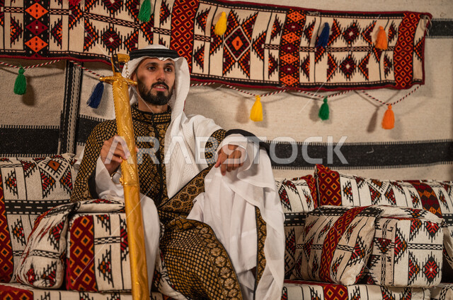 حب وشغف امتلاك السيوف التراثية, الجلوس داخل خيمة شعبية بإيماءات الاعتزاز والفخر, رجل عربي خليجي سعودي يرتدي الدقلة والغترة و يحمل السيف الى جانبه, التباهي بالأسلحة التقليدية السعودية, توارث العادات و التقاليد الأصيلة