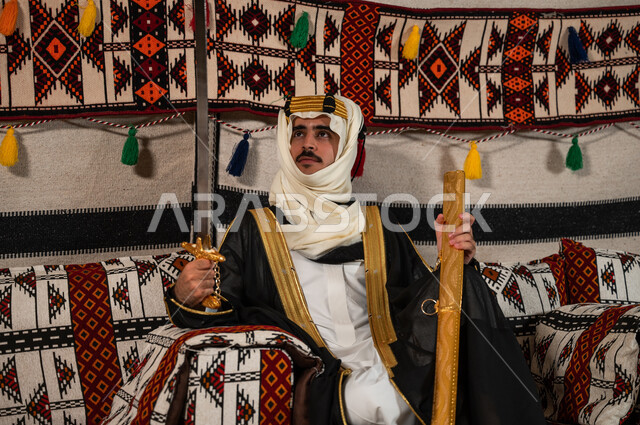 Wearing popular costumes on the anniversary of the founding of 1727, the day of the founding of the first Saudi state, February 22, sitting and looking at the sword with gestures of pride and honor, a Saudi Gulf Arab man wearing a bisht, a ghutra, and a golden headband and holding the Saudi sword, recalling the heroism of our ancestors and the inherited love of swords.