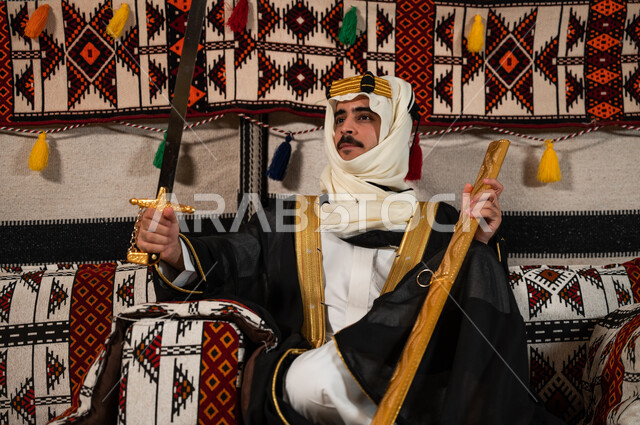 Sitting and looking at the sword with gestures of pride and honor, a Saudi Gulf Arab man wearing the bisht, ghutra, and golden headband and holding the Saudi sword, recalling the heroism of the ancestors and the inheritance of the love of swords, wearing popular costumes on the anniversary of the founding 1727, the day of the founding of the first Saudi state, February 22
