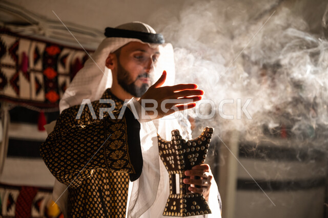 روائح عطرية محببة لدى أبناء الخليج, أصالة العادات والتقاليد السعودية, صورة مقربة لرجل عربي خليجي سعودي يرتدي الغترة والدقلة يقوم بالتبخر والتطيب بالبخور باستخدام المبخرة, استخدام خشب العود في المناسبات السعودية