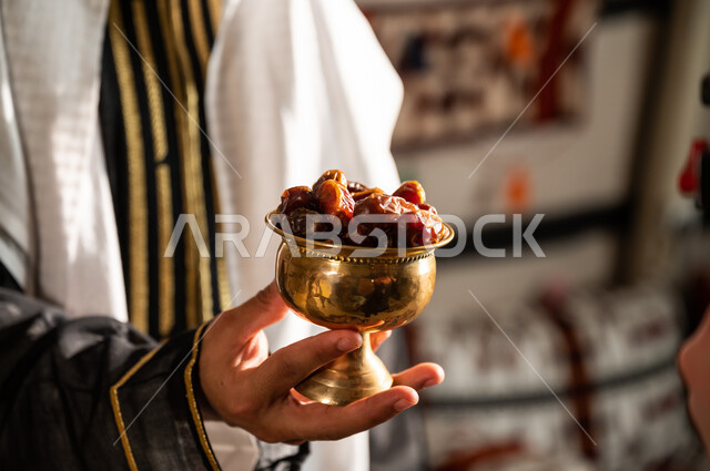 عادات وتقاليد سعودية شعبية في إكرام الضيف بتقديم المنتجات الوطنية الزراعية, طبق نحاسي مليء بالتمور، محاصيل محلية سعودية، صورة مقربة ليد رجل عربي سعودي خليجي يرتدي الغترة والدقلة يحمل بيده تمريه ذهبية, حسن الضيافة لدى السعوديين