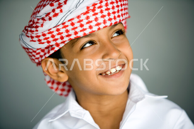 ملامح طفولية ناعمة وبريئة ، بورتريه مقرب لوجه ولد عربي  خليجي إماراتي يبتسم يرتدي العمامة الحمراء والكندورة التقليدية وينظر نحو الأعلى  ,  نظرات مليئة بالفرح والمتعة   ، خلفية رمادية .