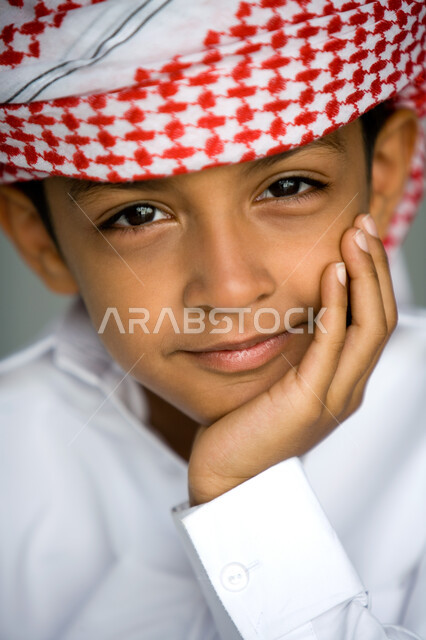 ملامح طفولية بريئة ، بورتريه مقرب لوجه ولد عربي  خليجي إماراتي يبتسم يرتدي العمامة الحمراء والكندورة التقليدية يضع يده تحت رأسه وينظر إلى الكاميرا  ,  الشعور بالسعادة والسرور  ، خلفية رمادية .