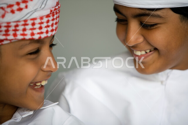 ملامح طفولية بريئة ، بورتريه مقرب لوجه ولدين عربيين  خليجيين إماراتيين  مبتسمين يرتديان العمامة الحمراء والكندورة التقليدية وينظران إلى بعضهما ,  تعابير الضحك والاستمتاع  ، قضاء الوقت  مع الأصدقاء بالمرح والتسلية ، خلفية رمادية .