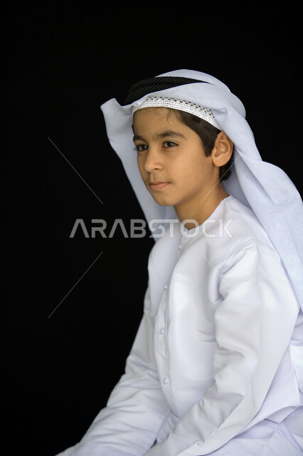 ملامح طفولية بريئة  تتسم بالأناقة والوسامة  ، بورتريه مقرب لصبي عربي  خليجي إماراتي  يرتدي العمامة البيضاء والكندورة التقليدية وينظر إلى الأمام ,  إيماءات وجه تدل على التركيز والتخطيط  ، خلفية رمادية .