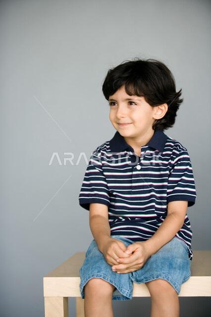 نظرات طفولية  تتسم بالأناقة والبراءة  والخجل ، بورتريه لصبي عربي  خليجي إماراتي مبتسم يرتدي الزي الكاجوال وينظر إلى الجانب , الجلوس على مقعد خشبي  ، خلفية رمادية .