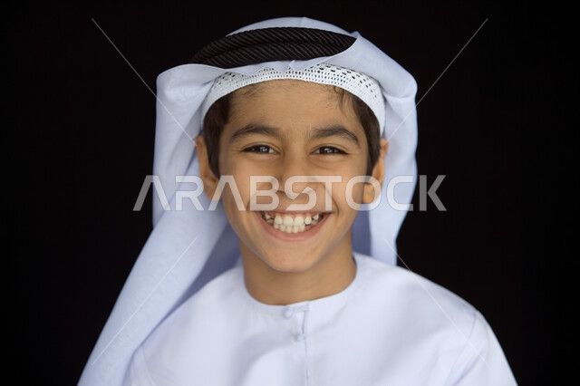ضحكة مشاكسة مليئة بالفرح  ، بورتريه مقرب لوجه صبي عربي  خليجي إماراتي مبتسم يرتدي العمامة البيضاء والغترة  والكندورة التقليدية وينظر إلى الكاميرا ,  ملامح طفولية بريئة  تتسم بالأناقة والوسامة ، خلفية رمادية .