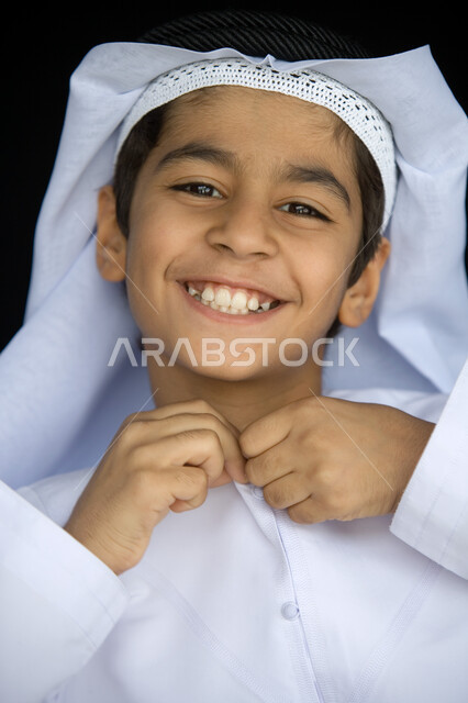 الاهتمام بالأناقة والمظهر الخارجي   ، بورتريه مقرب لصبي عربي  خليجي إماراتي مبتسم يرتدي العمامة البيضاء ويقوم بتزرير الكندورة التقليدية وينظر إلى الكاميرا , ضحكة طفولية مليئة بالسعادة ، خلفية رمادية .