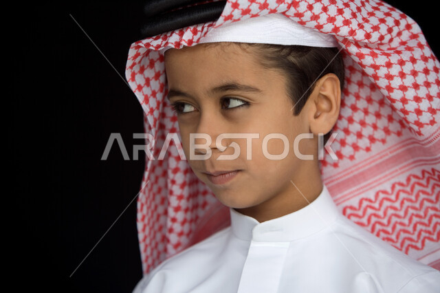 إيماءات وجه تدل على التركيز والتخطيط  ، بورتريه مقرب لصبي عربي  خليجي إماراتي  يرتدي العمامة الحمراء والعقال والكندورة التقليدية وينظر إلى الجانب بتمعن , ملامح طفولية  تتسم بالذكاء والفطنة  ، خلفية سوداء .
