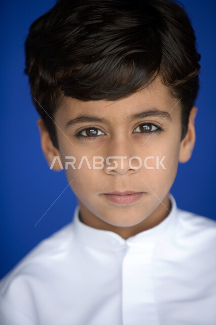 نظرات طفولية  تتسم بالتحديق والتأمل  ، بورتريه مقرب لوجه صبي عربي  خليجي إماراتي يبتسم   يرتدي الكندورة التقليدية وينظر إلى الكاميرا ,  مظهر طفولي يتصف بالأناقة والوسامة  ، خلفية زرقاء .