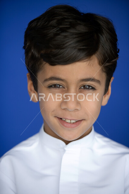 مظهر طفولي يتصف بالأناقة والوسامة  ، بورتريه مقرب لوجه صبي عربي  خليجي إماراتي يبتسم   يرتدي الكندورة التقليدية وينظر إلى الكاميرا ,  نظرات طفولية  تتسم بالتخطيط والتفكير  ،  خلفية زرقاء .