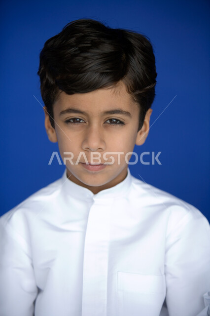 نظرات طفولية  تتسم بالتخطيط والتفكير  ، بورتريه مقرب لوجه صبي عربي  خليجي إماراتي يبتسم   يرتدي الكندورة التقليدية وينظر إلى الكاميرا ,  مظهر طفولي يتصف بالأناقة والوسامة  ، خلفية زرقاء .